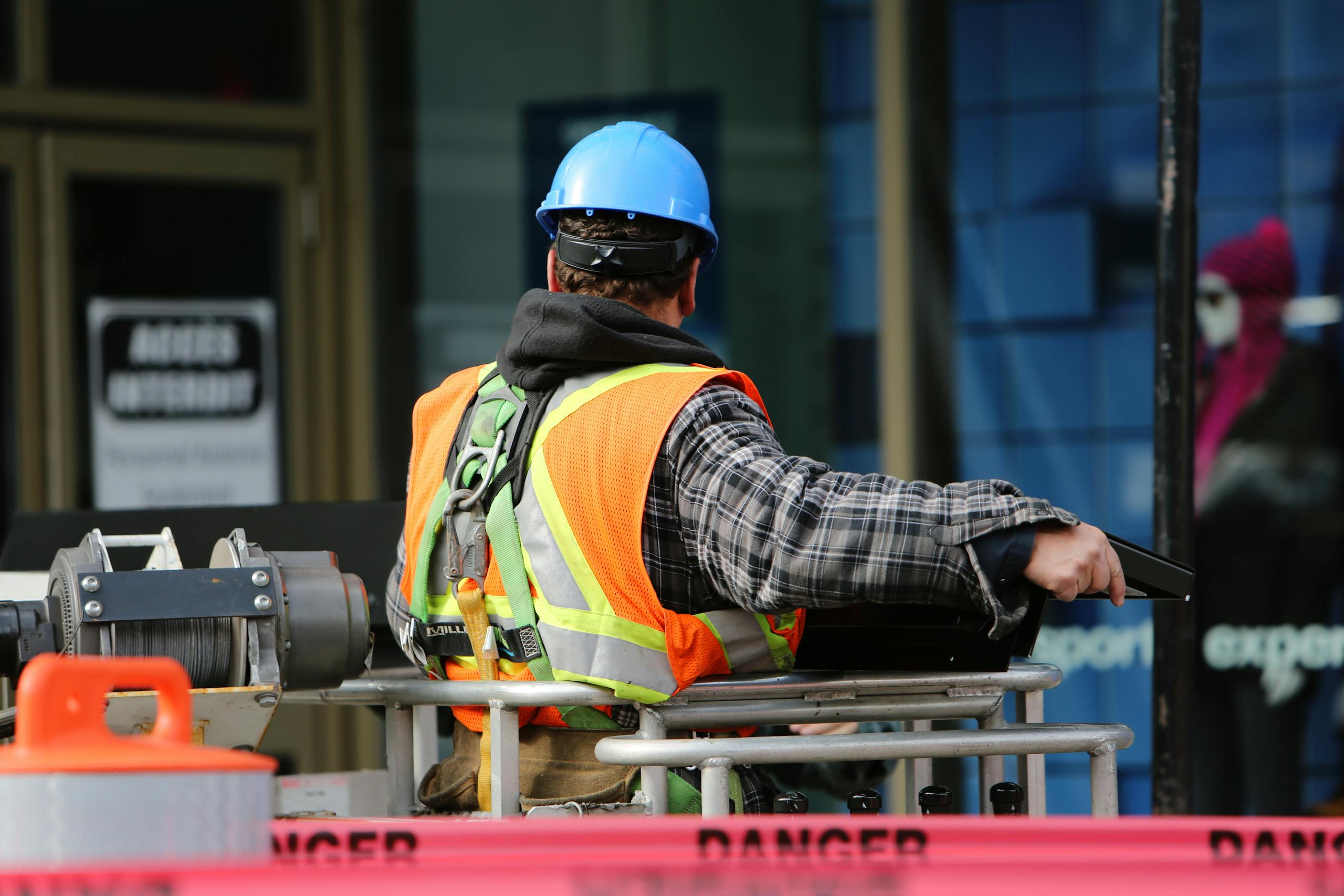 A construction worker wearing a blue helmet and safety vest operates equipment at an urban construction site.