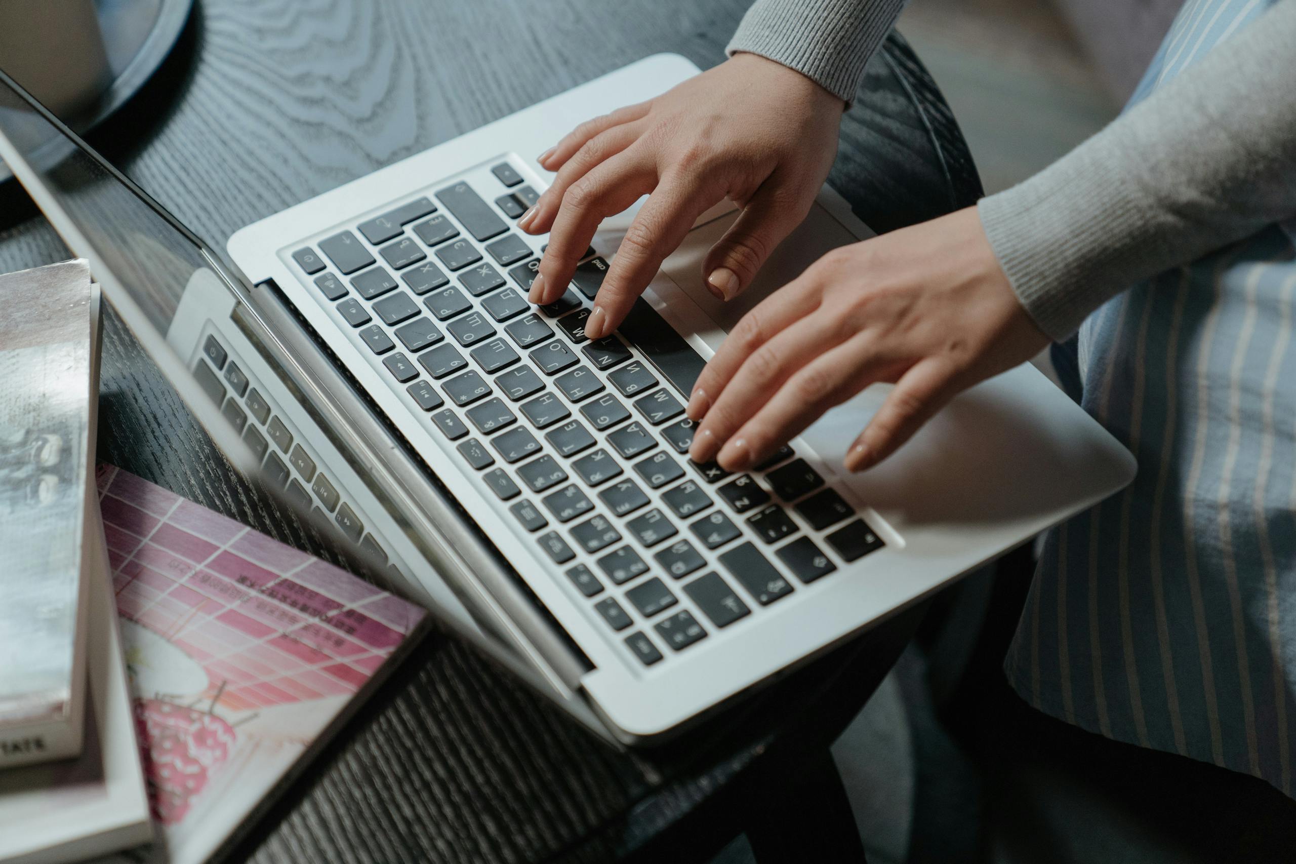 Close-up of a woman's hands typing on a laptop indoors, with books on the table.