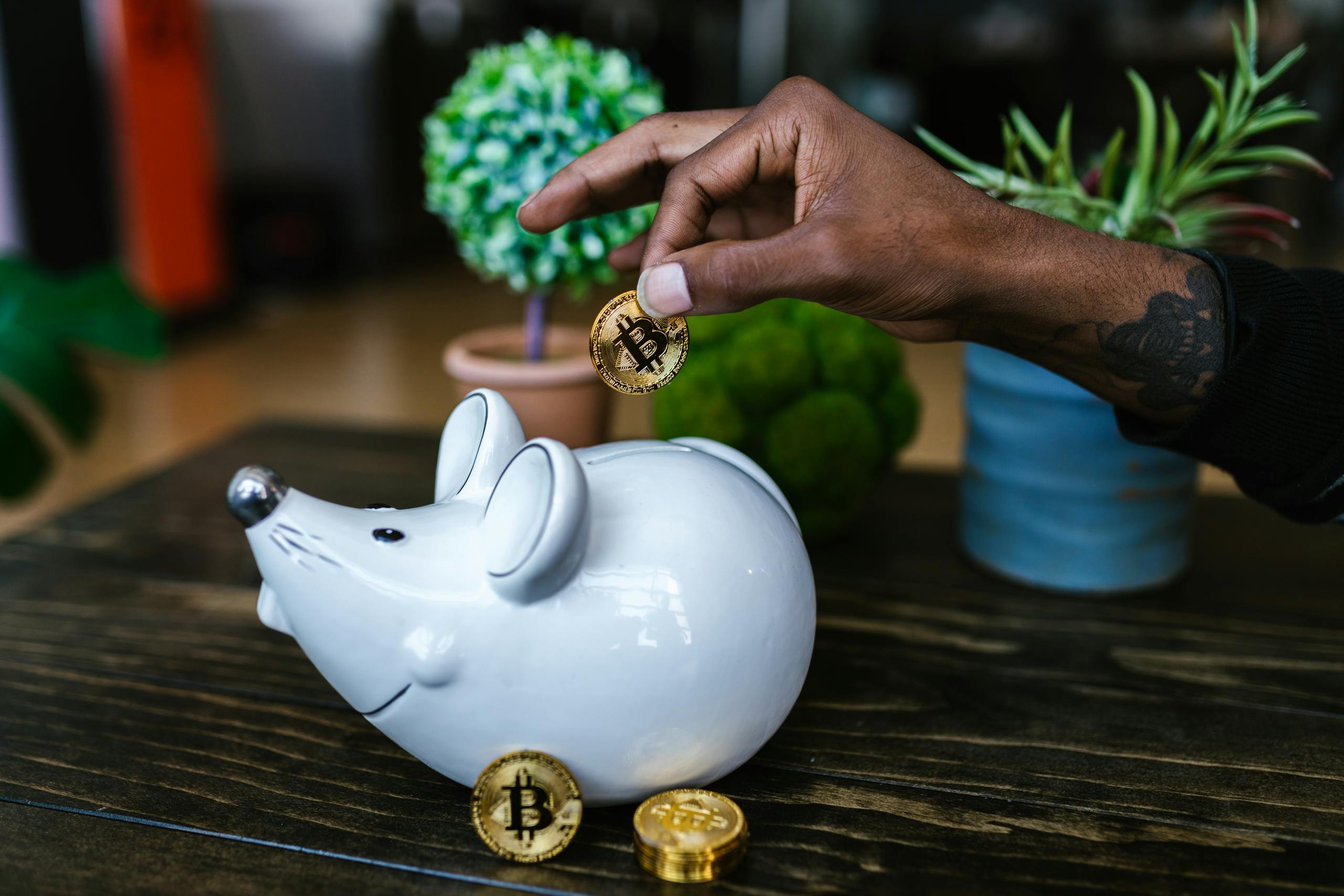 Close-up of hand placing bitcoin into a mouse-shaped piggy bank on a table.