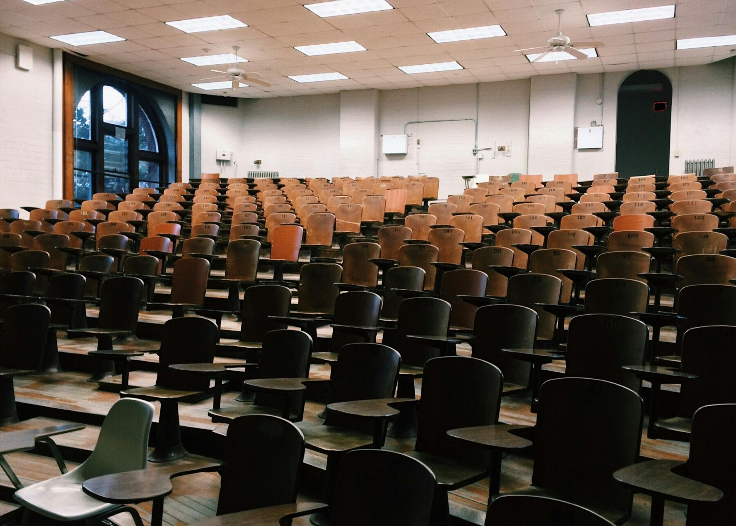 Empty university lecture hall with wooden chairs and large windows.